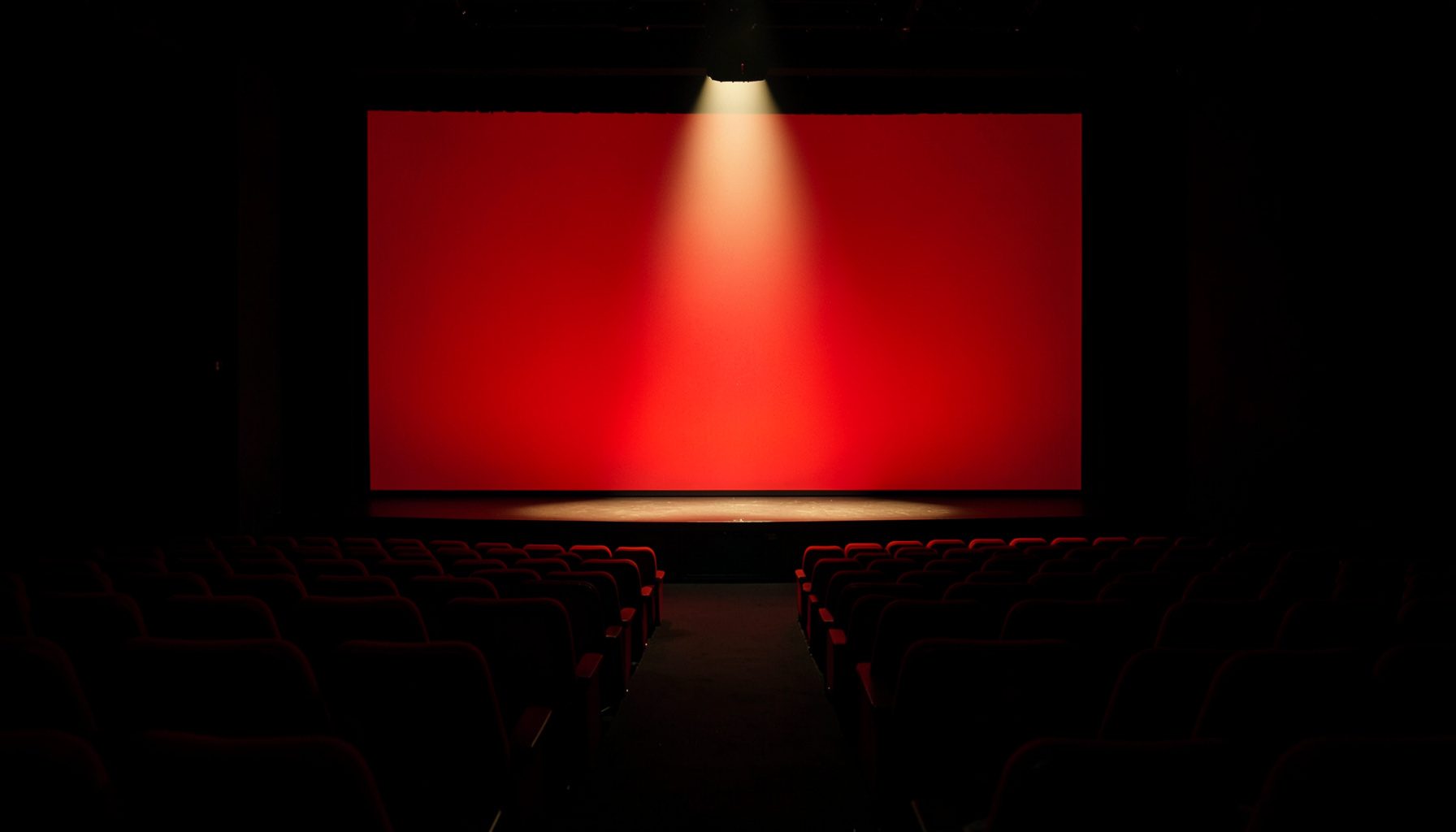 Darkened cinema auditorium with rows of red seats fading into shadow, a single warm spotlight on the stage